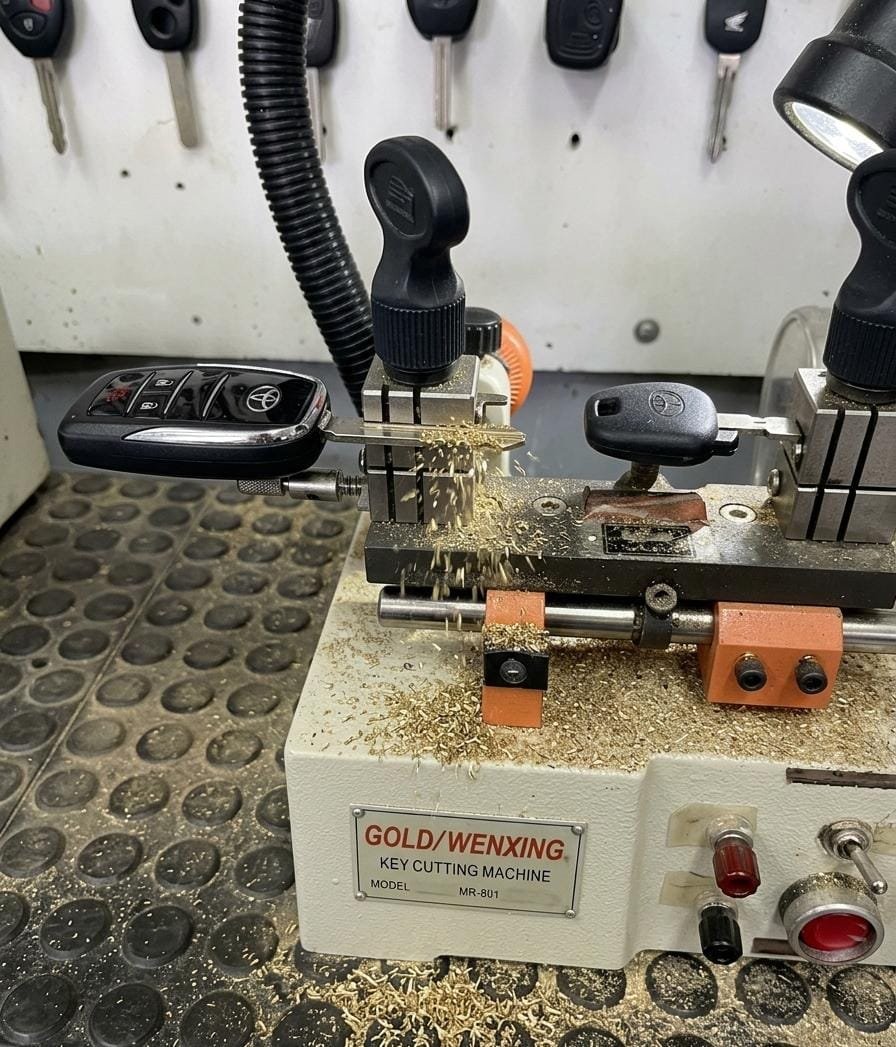 Mão de chaveiro trabalhando em máquina de cópia de chaves automotivas com veículo ao fundo em oficina profissional